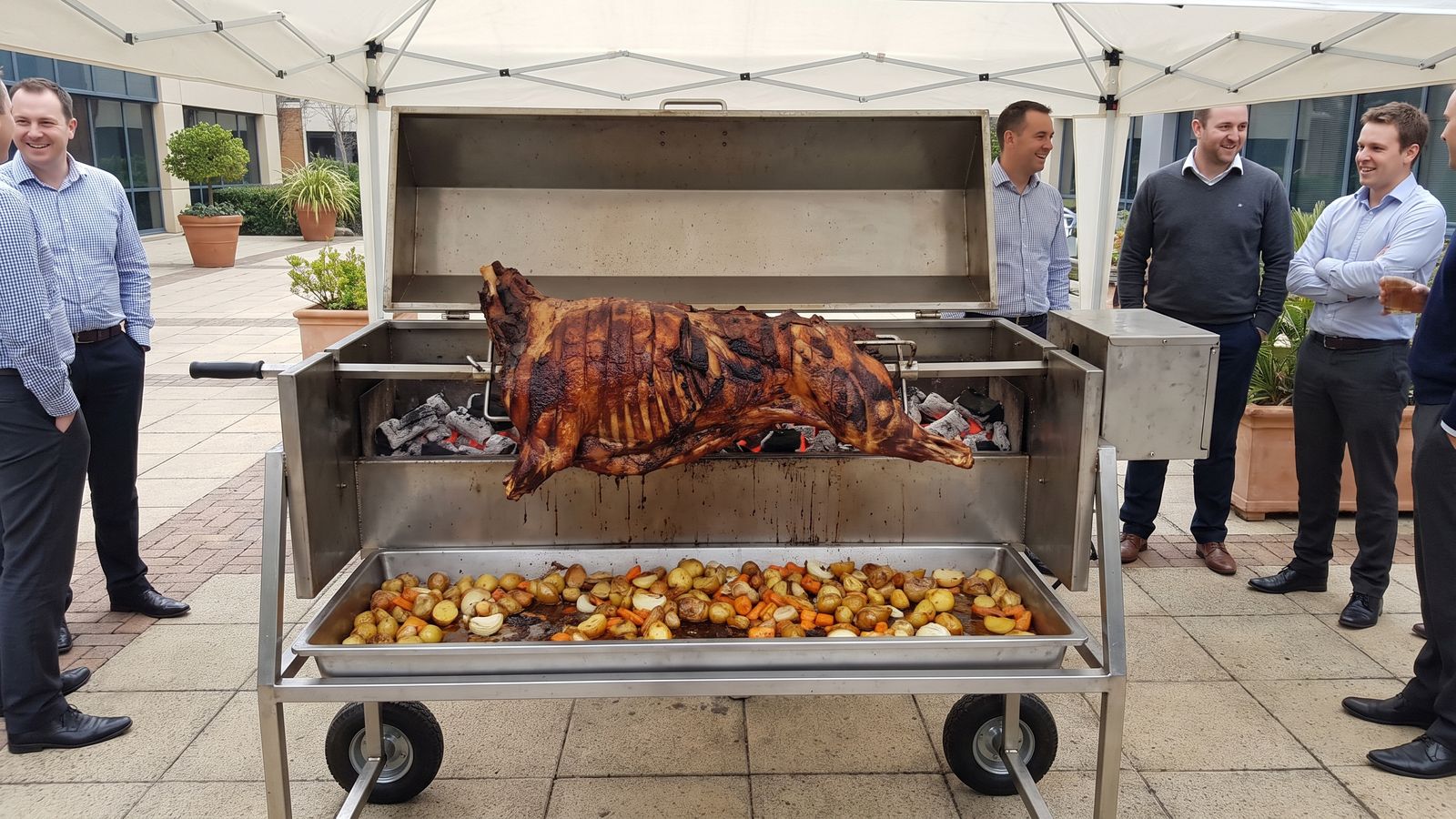 Whole lamb spit braai on a stainless machine in a paved office-park courtyard, the kind Spit Braai Feast caters in Kuils River