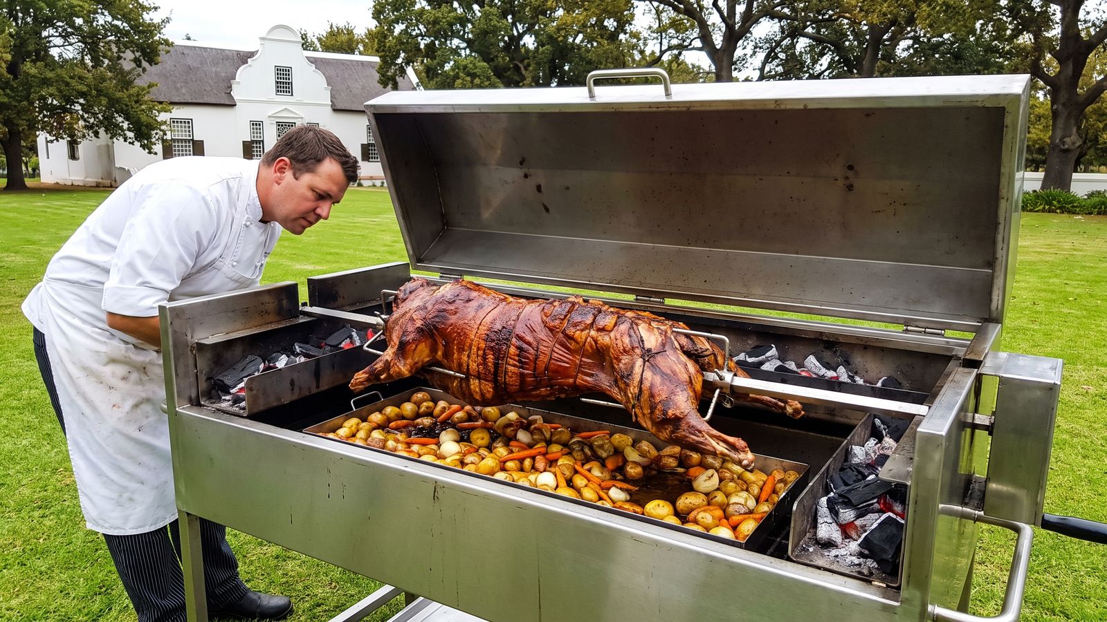 Whole lamb spit braai on a stainless machine on a farm lawn with a gabled farmhouse behind, the kind of venue Spit Braai Feast caters in Stellenbosch