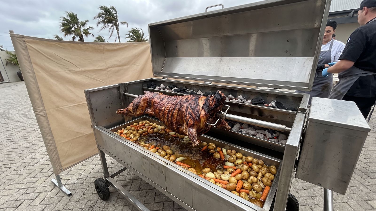 Whole lamb spit braai on a stainless machine beside a canvas windbreak in a paved courtyard, the set-up Spit Braai Feast uses in Strand