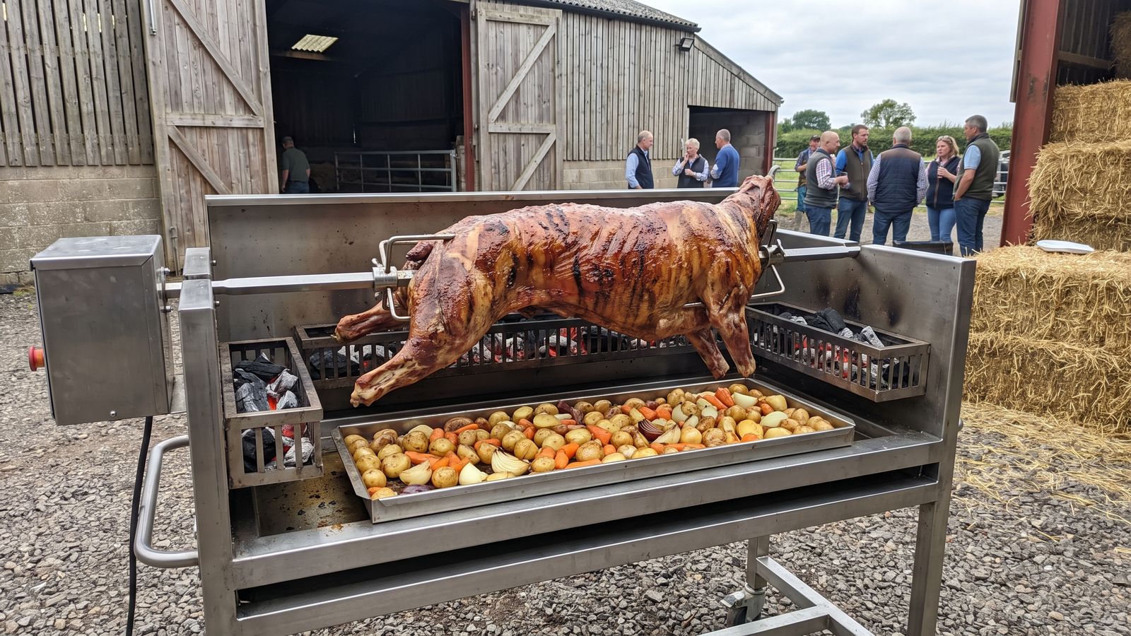 Whole lamb spit braai on a stainless machine on a gravel yard outside a barn venue, the kind Spit Braai Feast cooks in Wellington